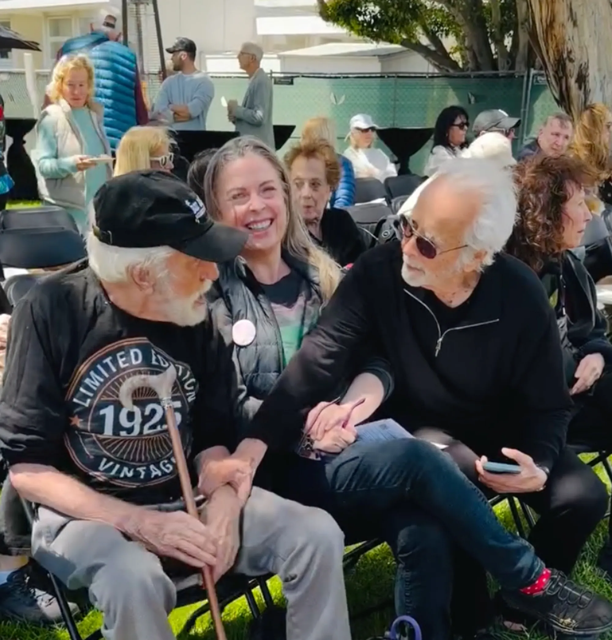 Two men, one with a white beard, sitting outdoors with other people and holding hands.