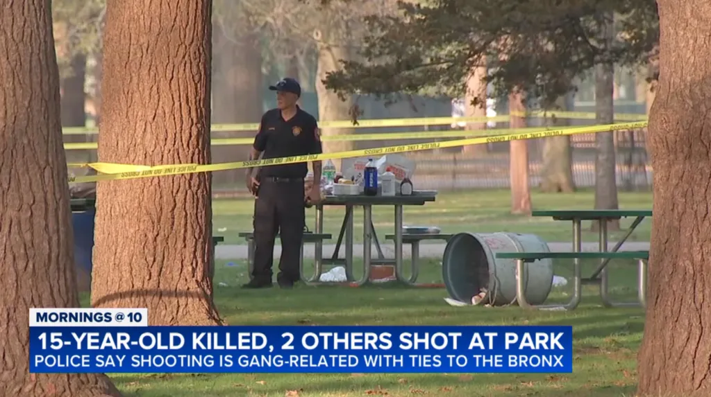 A police officer standing behind "Police Line Do Not Cross" tape at a park with picnic tables.