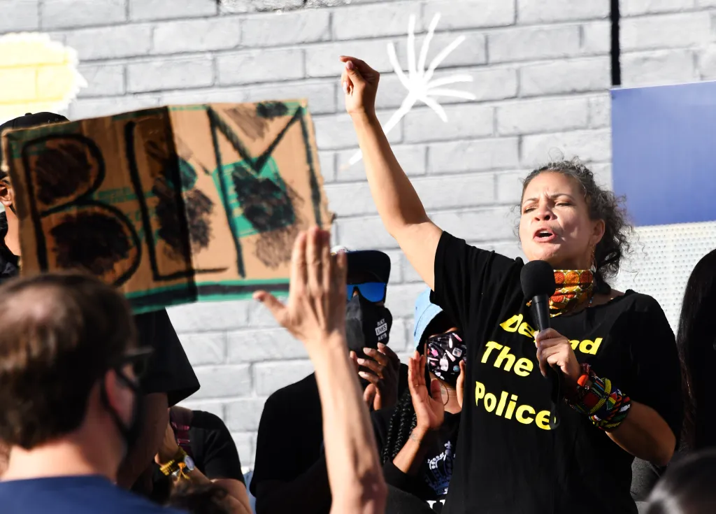 Dr. Melina Abdullah speaking into a microphone at a Black Lives Matter rally, with a raised fist and a sign that says "BLM" in the foreground.