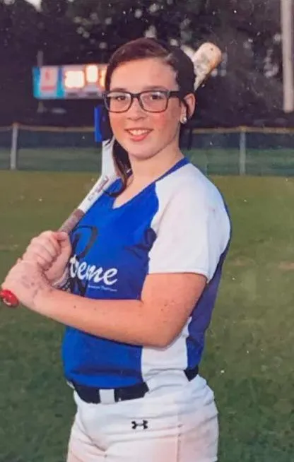 17-year-old Keziah Arionna Luker, wearing a blue and white softball uniform and glasses, holding a baseball bat.