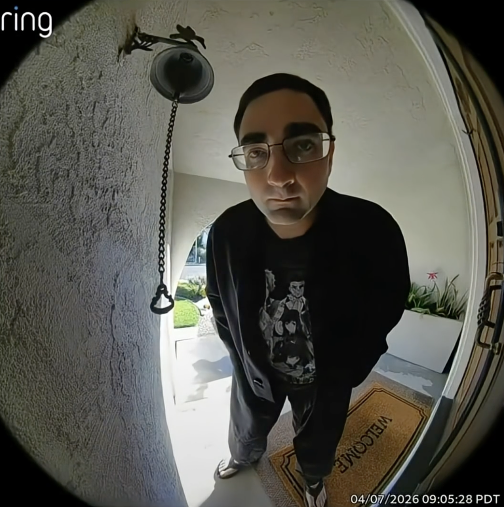 A man in glasses, a black sweatshirt, and dark pants, stands on a "Welcome" mat, looking directly into a Ring doorbell camera.