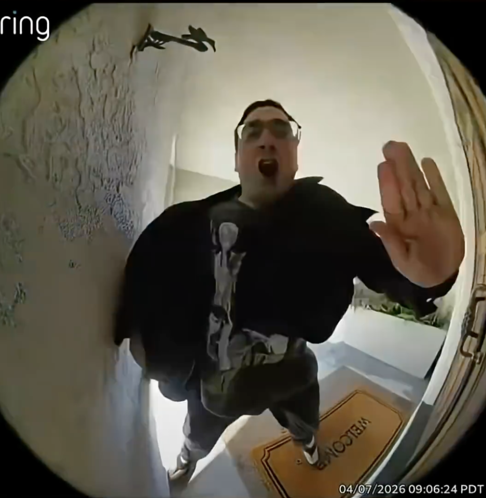 A man with glasses and a black jacket, captured by a Ring doorbell camera, standing on a "WELCOME" mat, with his mouth open in a shout and one hand extended forward.