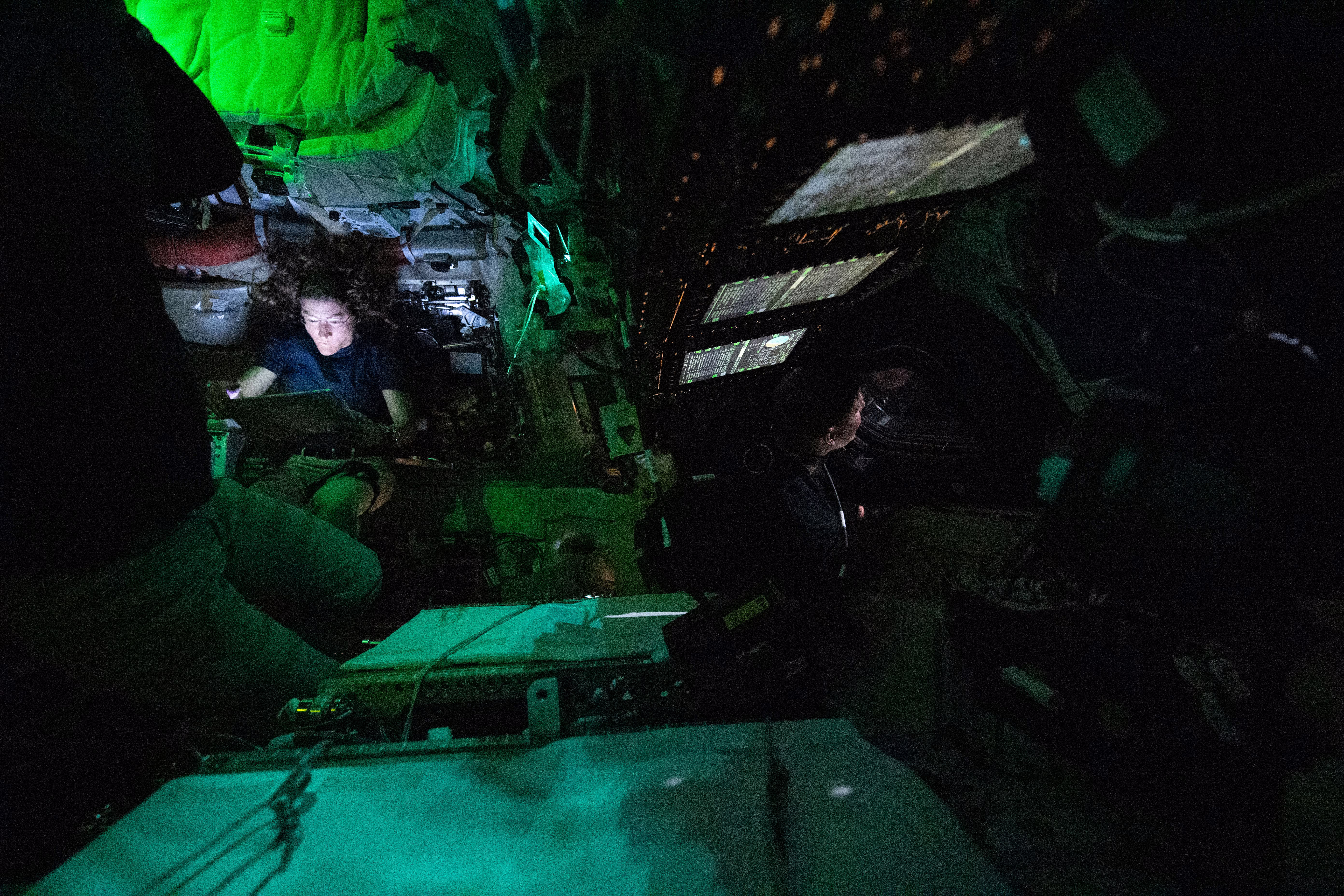 The mostly-dark interior of a small spacecraft, illuminated by various monitor screens. Two astronauts can be seen, one looking out the window, another looking at a laptop.