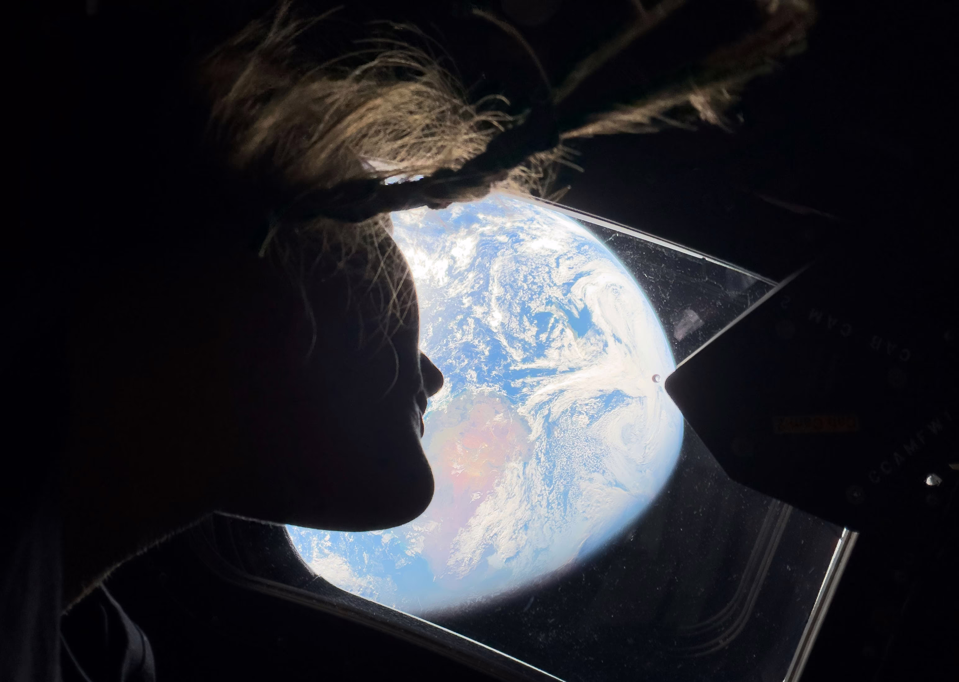 An astronaut, seen in profile, with the brightly-lit earth in the background, seen through a window
