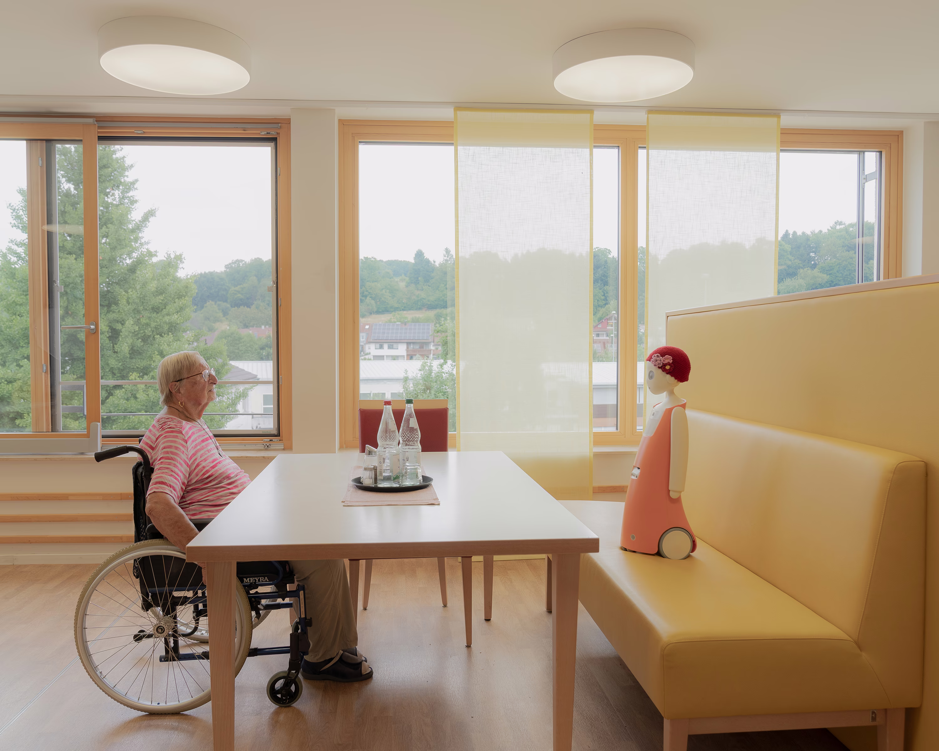 A person in a wheelchair sits at a table across from a small humanoid robot.