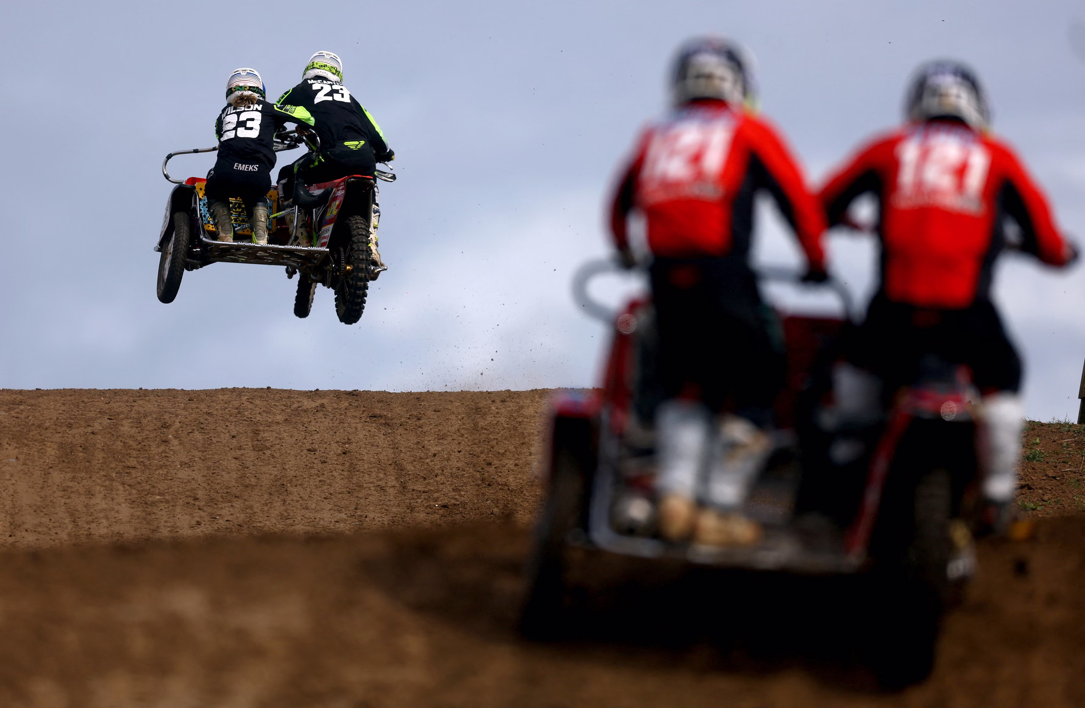 Racers on motorcycle-and-sidecar rigs leap over hills on a dirt track.