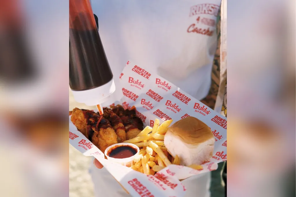 A person squeezes hot sauce from a bottle onto fried chicken served with fries, a bun, and a small container of sauce, all in a paper tray.
