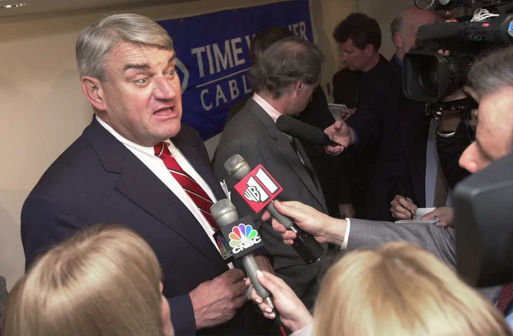 Time Warner Cable CEO Joseph Collins speaks at a news conference, surrounded by microphones from CNBC and WB.
