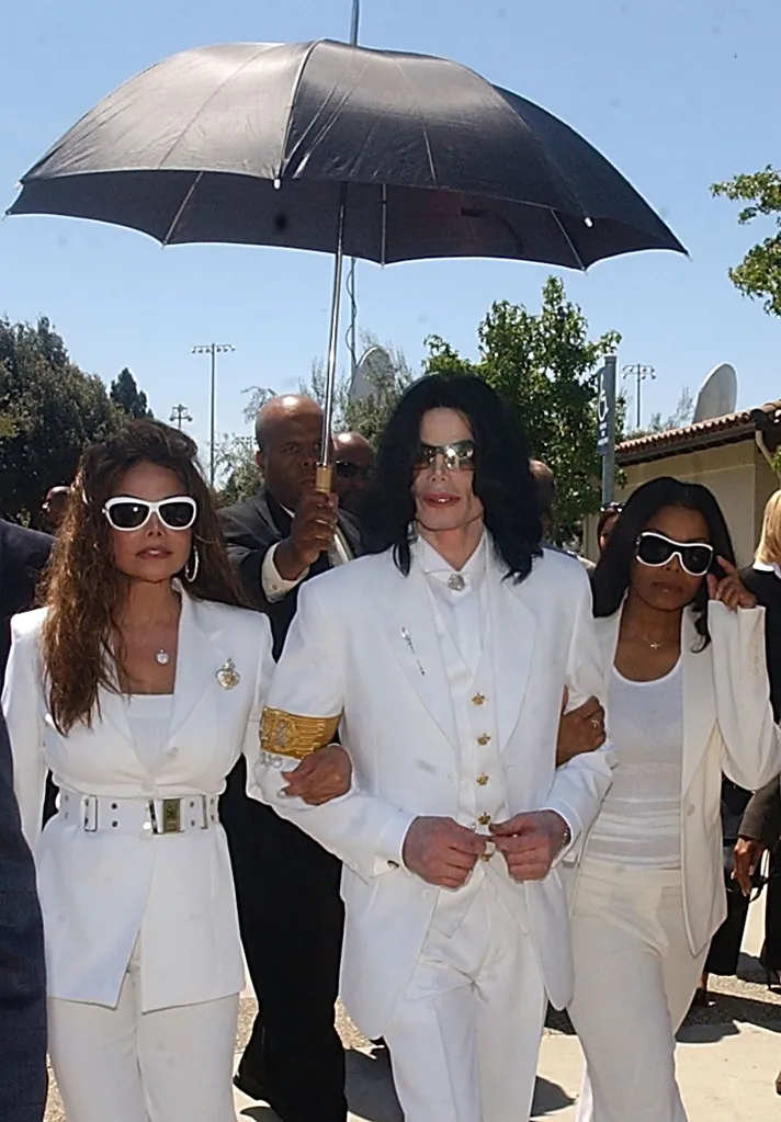 Michael Jackson, Janet Jackson, and La Toya Jackson in white suits and sunglasses, walking under a black umbrella.