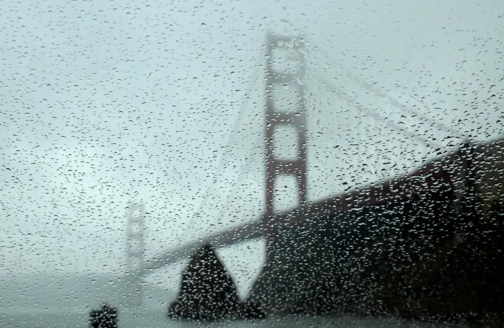 Raindrops bead on a car window with a blurred view of the Golden Gate Bridge in the background.