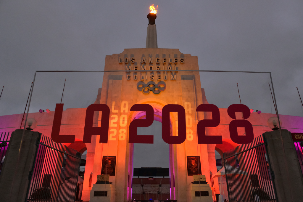 The Olympic cauldron atop the Los Angeles Memorial Coliseum burns, with a large "LA 2028" sign in front, illuminated by colored lights.
