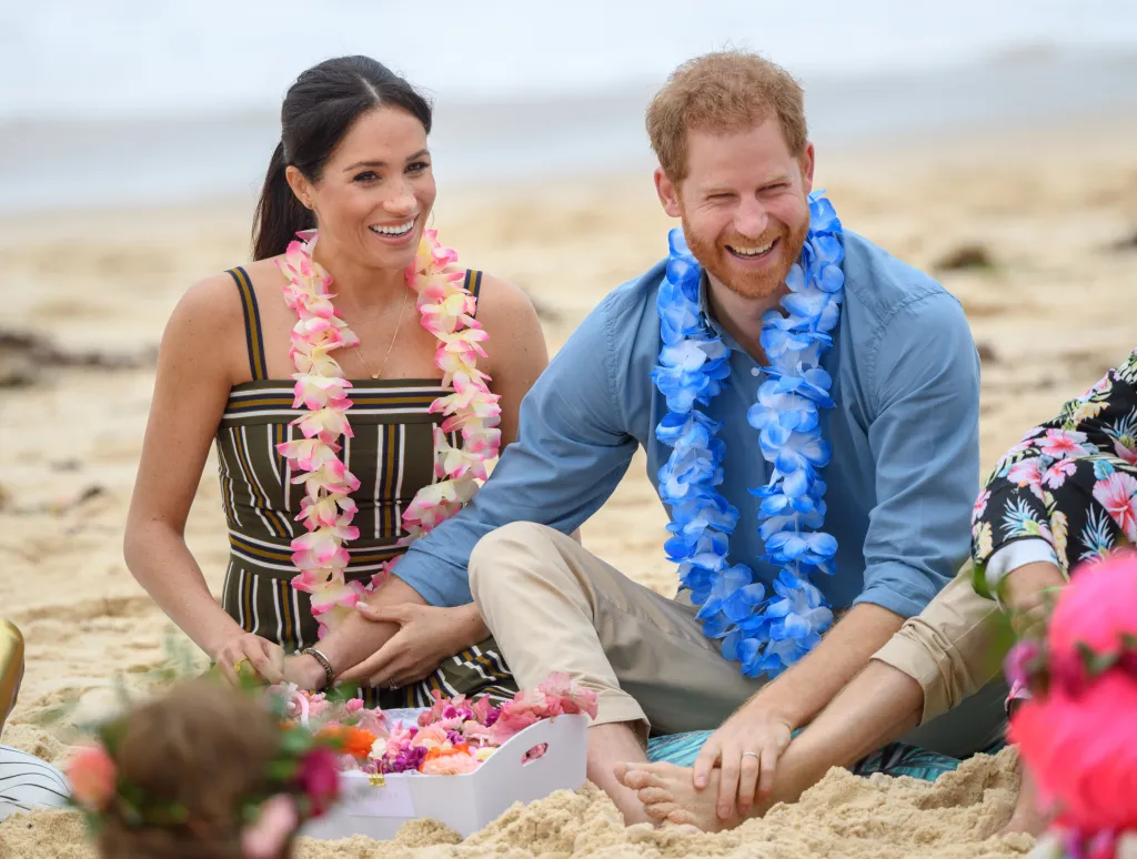 Prince Harry, in a blue shirt and khaki pants and blue flower lei, and Meghan Markle, in a black and white striped dress and a pink lei, the Duke and Duchess of Sussex, laughing while sitting on Bondi Beach wearing leis.