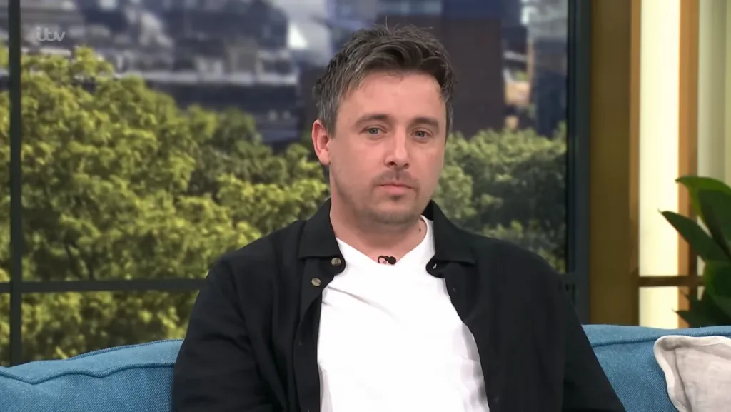 Steven Hamill, a 26-year-old male, wearing a black shirt over a white t-shirt, sits on a blue couch, looking directly into the camera.