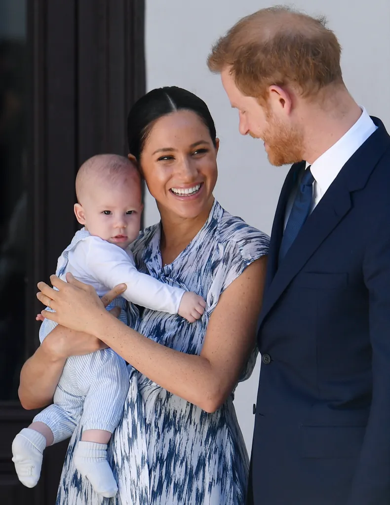 Meghan Markle smiles while holding baby Archie next to Prince Harry, who is looking down at his son.