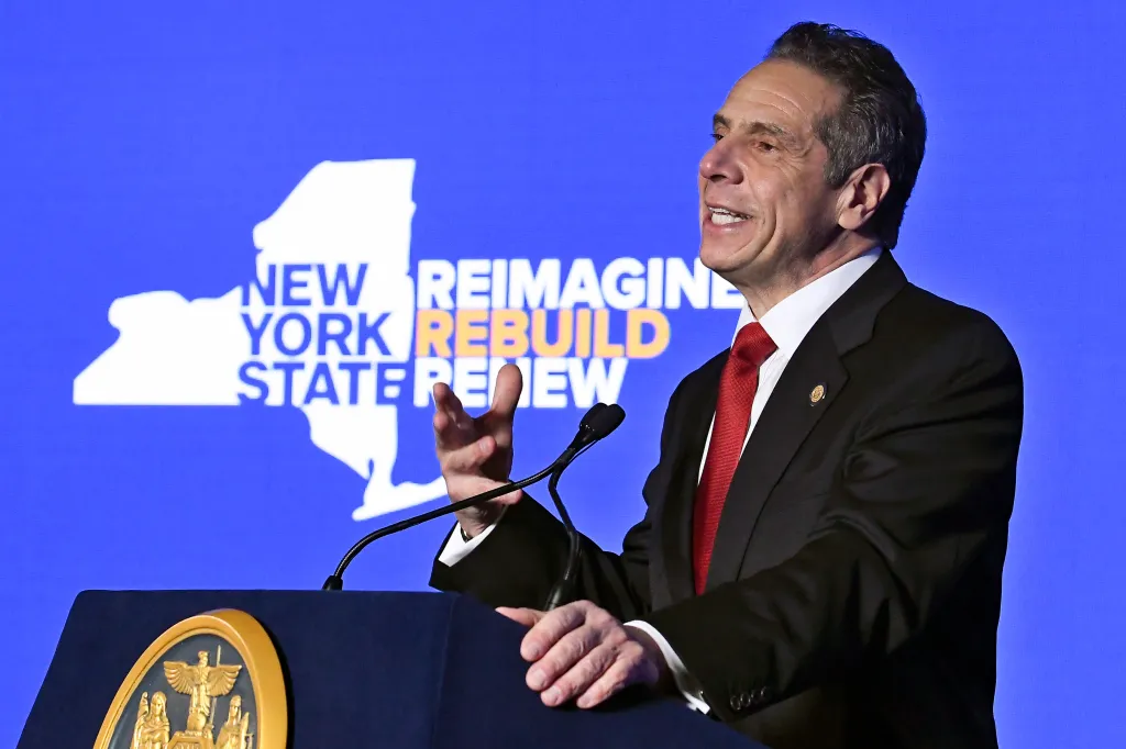 New York Gov. Andrew Cuomo speaks at a podium with a blue screen behind him displaying the words "NEW YORK REIMAGINE REBUILD RENEW".
