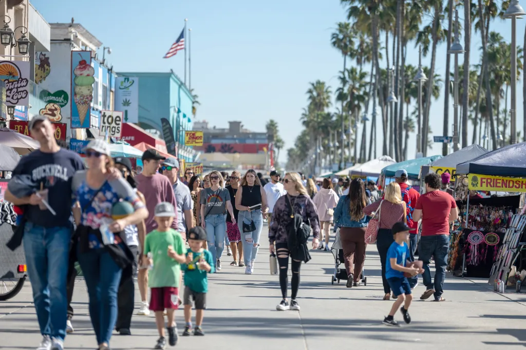Crowds of people walk along the Venice Beach boardwalk in warm weather.