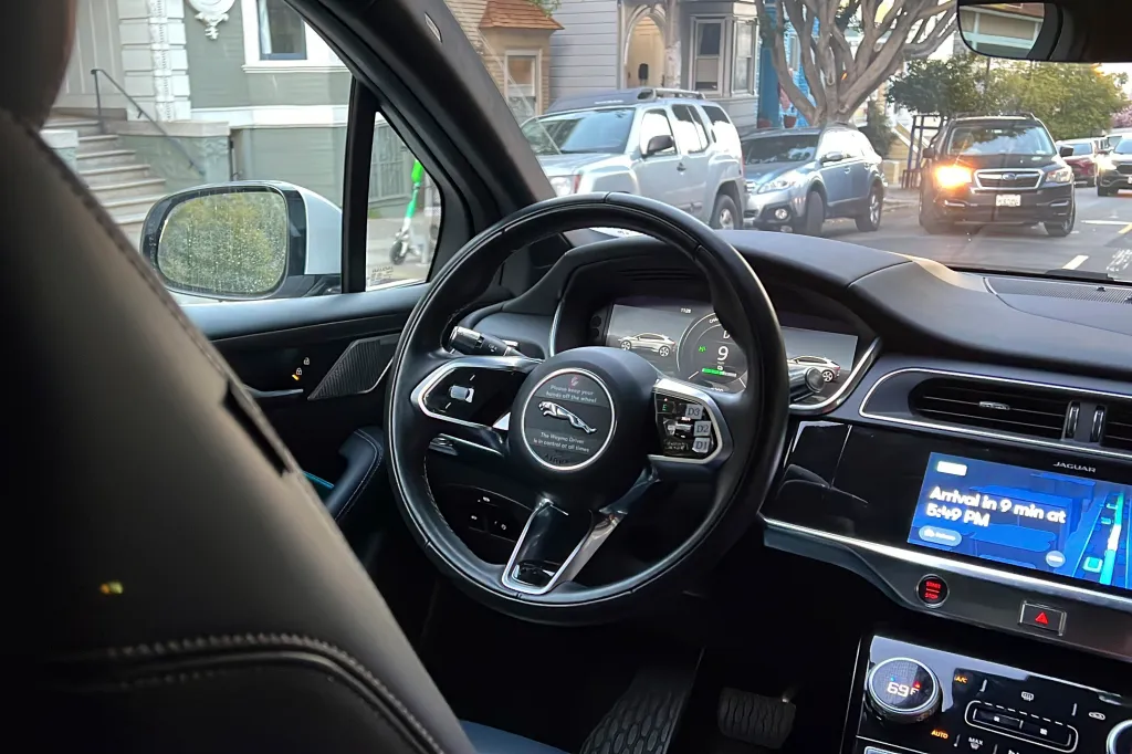 Interior view of a Waymo driverless taxi, showing the steering wheel with a "Please keep your hands off the wheel" message, and the dashboard displaying an arrival time.