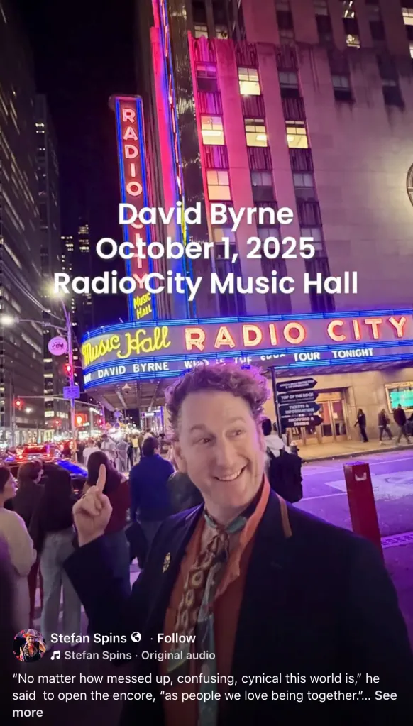 A man in a scarf points up at the bright neon sign of Radio City Music Hall in New York City, where text announces a "David Byrne October 1, 2025" concert.