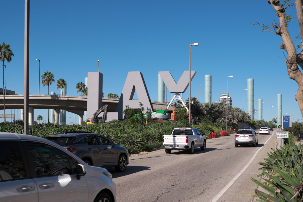 The iconic LAX sign, made of large gray letters, is seen from a road with cars driving by, while parts of the sign are being removed by construction equipment.