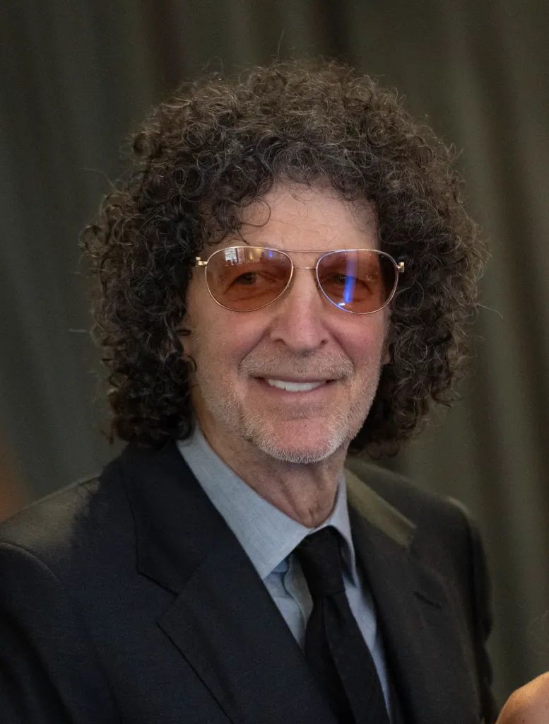 Howard Stern smiles, wearing a dark suit, light blue shirt, dark tie, and gold-rimmed aviator sunglasses with tinted lenses.