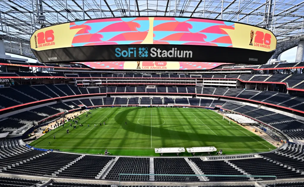 A wide view of the SoFi Stadium field with a new grass pitch, surrounded by empty black seats, and a large oval jumbotron overhead displaying FIFA World Cup 2026 Los Angeles branding.