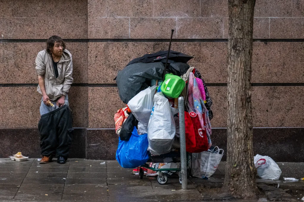 A homeless man stands on the street next to a cart filled with belongings.