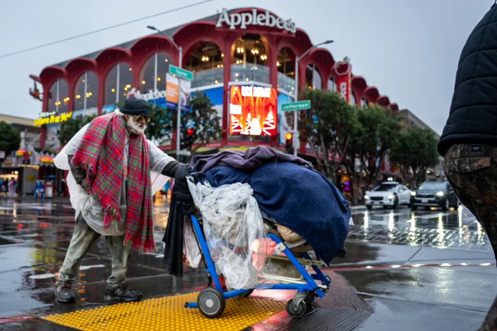 A homeless person pushing a handcart with belongings on a wet street in front of an Applebee's restaurant in San Francisco.