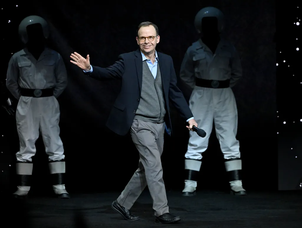 Rick Moranis waving to the audience with two costumed figures in the background.
