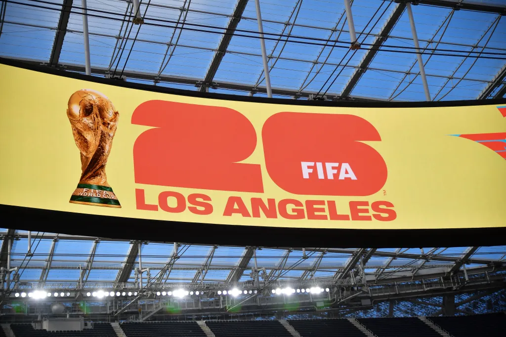World Cup 2026 branding on a large screen at SoFi Stadium, with the FIFA World Cup trophy displayed next to "26 FIFA Los Angeles" against a yellow background.