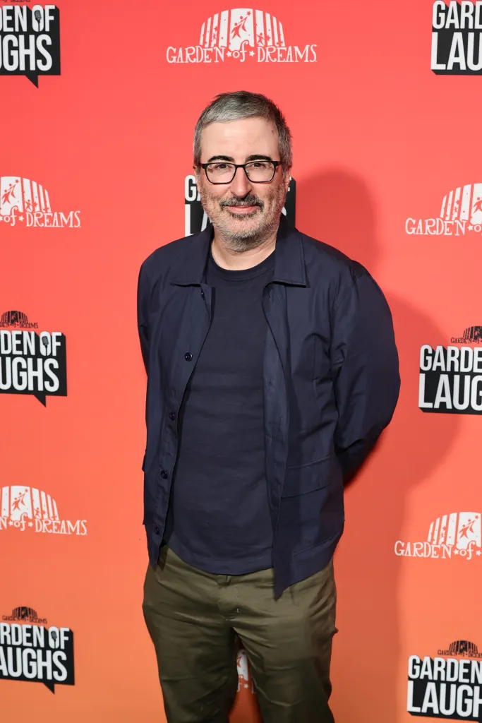 John Oliver smiling in front of a red backdrop with "Garden of Laughs" and "Garden of Dreams" logos.