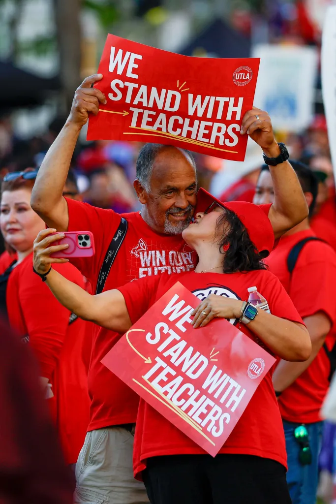 A man and woman at a rally holding signs that read "WE STAND WITH TEACHERS."
