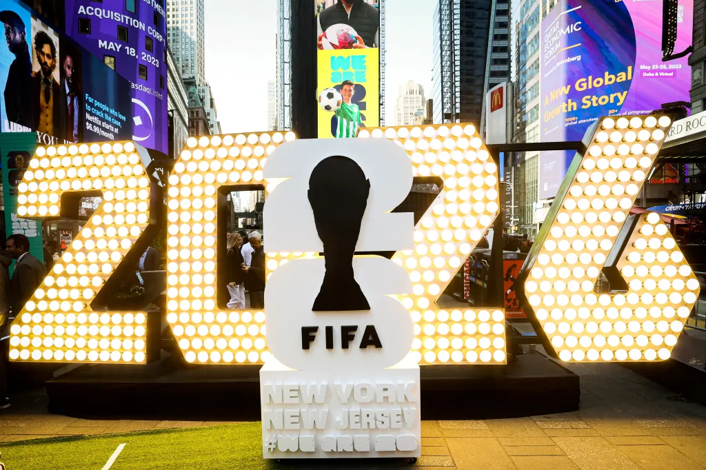 The FIFA World Cup 2026 logo, featuring the numbers 20 and 26 with a silhouette of the World Cup trophy as the "2" in 2026, is displayed in Times Square.