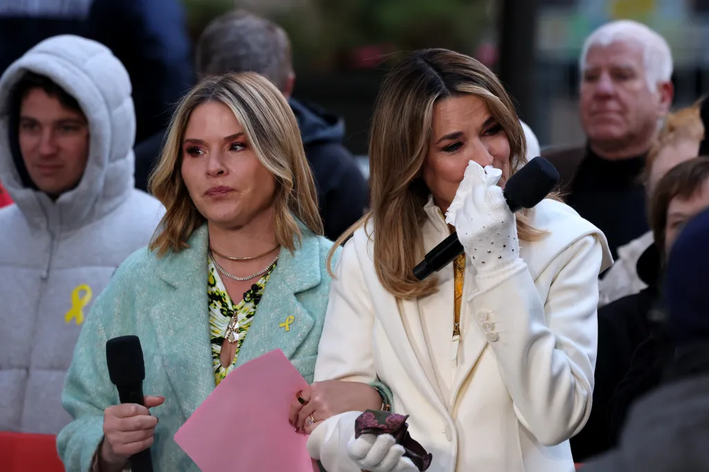 Savannah Guthrie, wearing a white coat and gloves, cries while holding a microphone, with a fellow "Today" show host beside her.