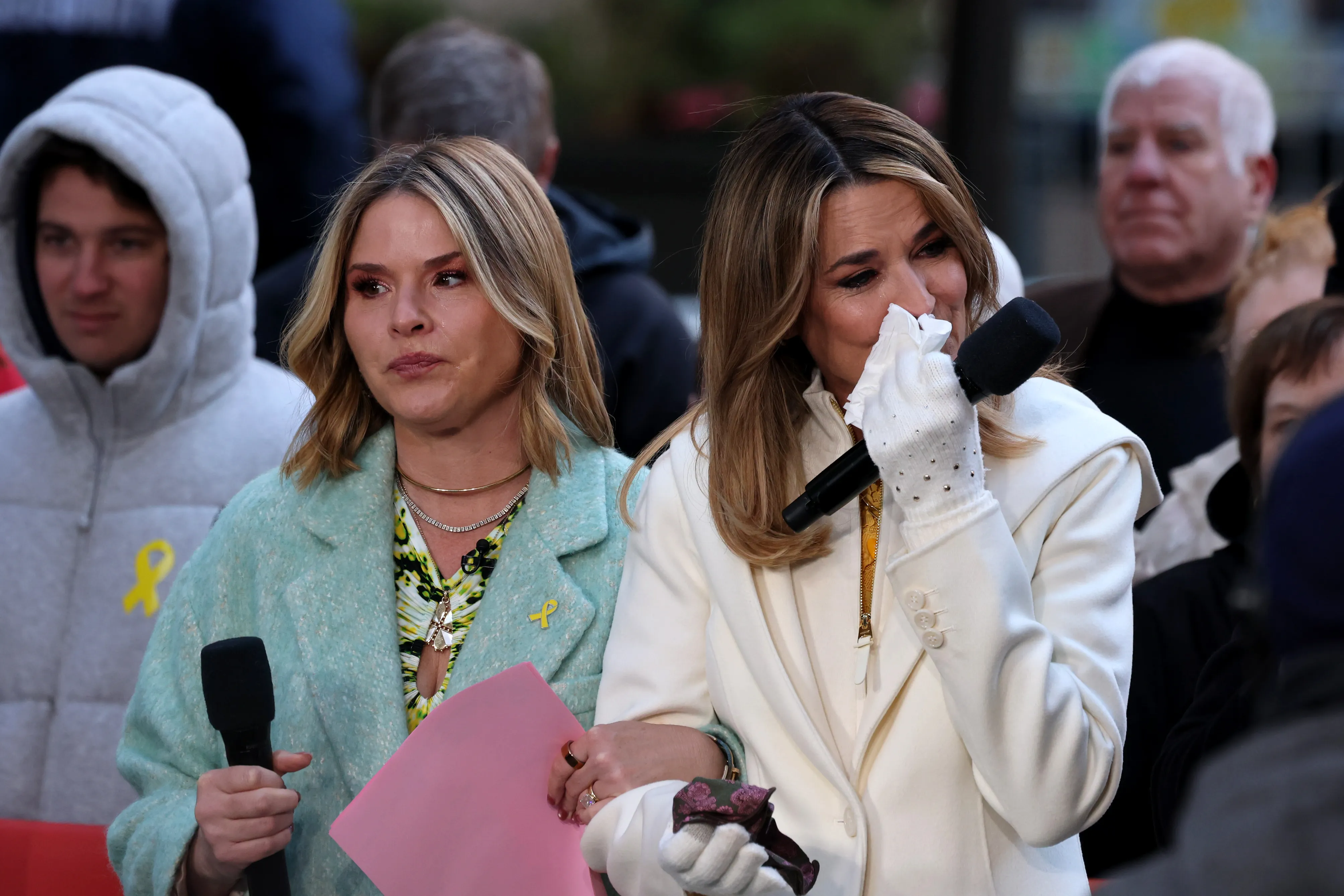 Savannah Guthrie crying into a tissue while holding a microphone, standing next to another woman, outside.