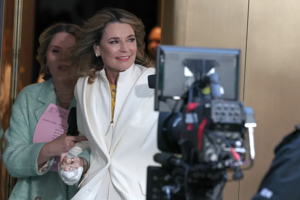 Savannah Guthrie returns to the Today show plaza in Manhattan, wearing a white jacket over a yellow shirt and smiling.