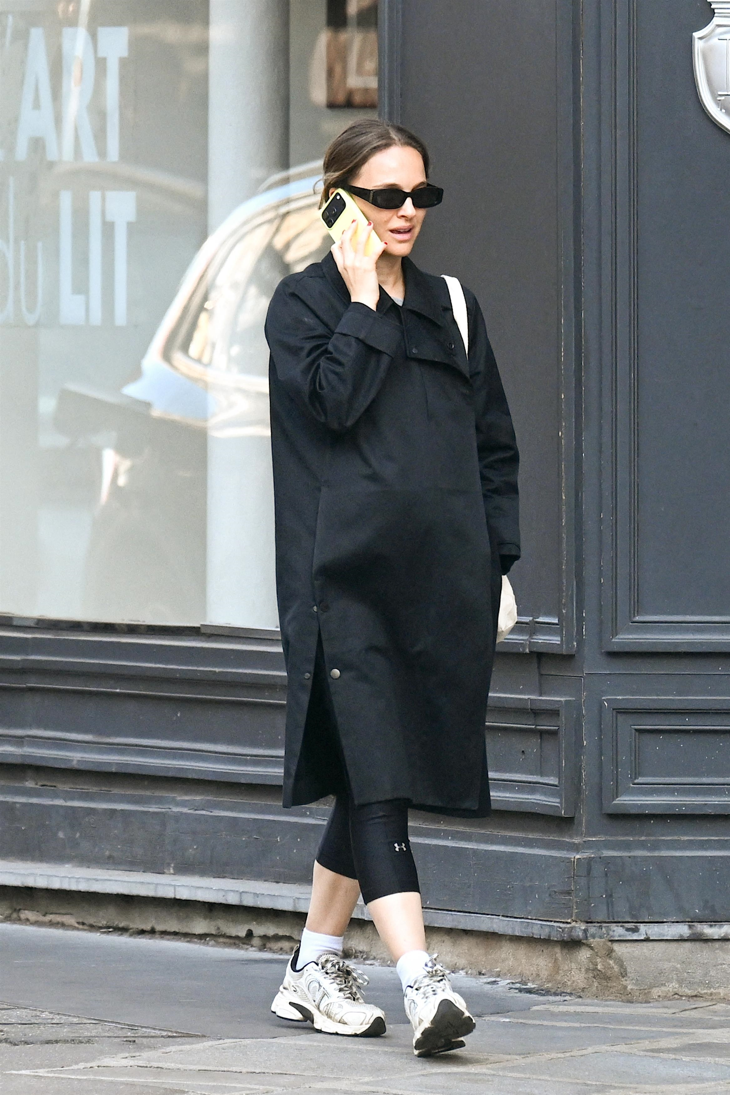 Natalie Portman in a black coat and black leggings, talking on a neon green phone, with a beige tote bag on her shoulder.