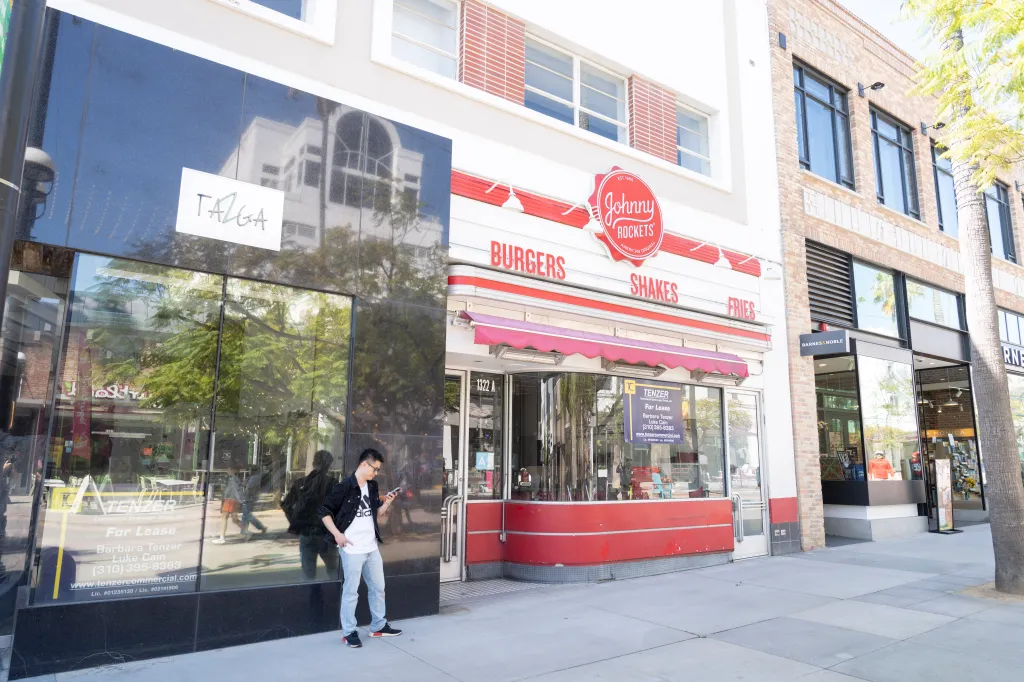 A man checks his phone outside Johnny Rockets on 3rd Street Promenade in Santa Monica, with a "For Lease" sign visible in the window next door.