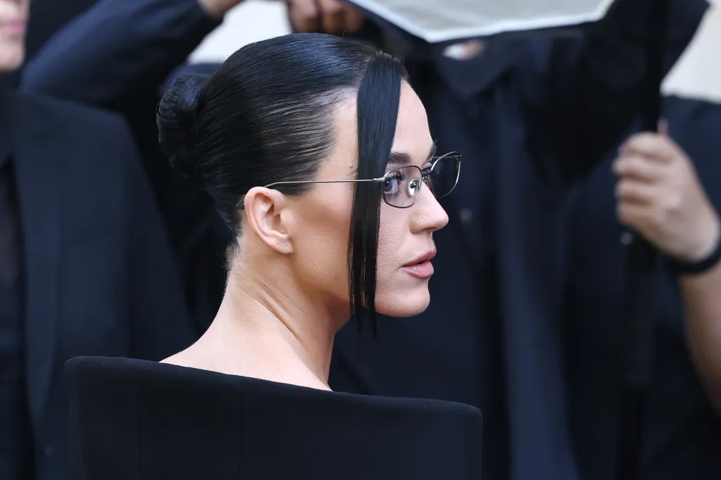 Katy Perry wearing glasses and a black gown, with her hair in a bun and two strands framing her face.