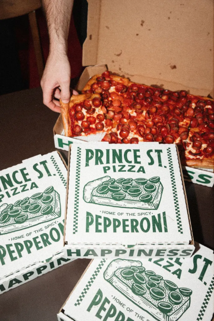 A person reaching into a box to pull out a slice of Prince Street Pizza's 