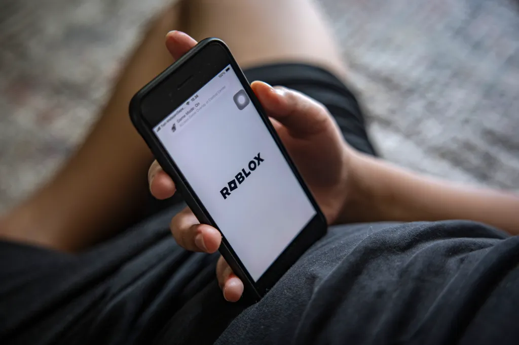 A 12-year-old boy holds a smartphone showing the Roblox app on its screen.