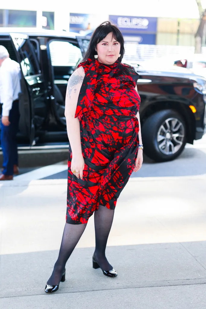 A woman with long dark hair, wearing a red and black patterned dress, black tights, and black heels, stands outside on a sunny day.