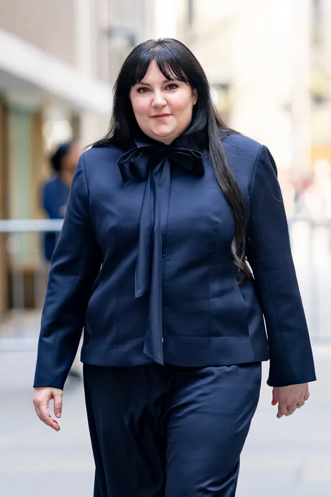 A woman with long dark hair, wearing a dark blue suit with a bow tied at the neck, walks forward and smiles slightly.