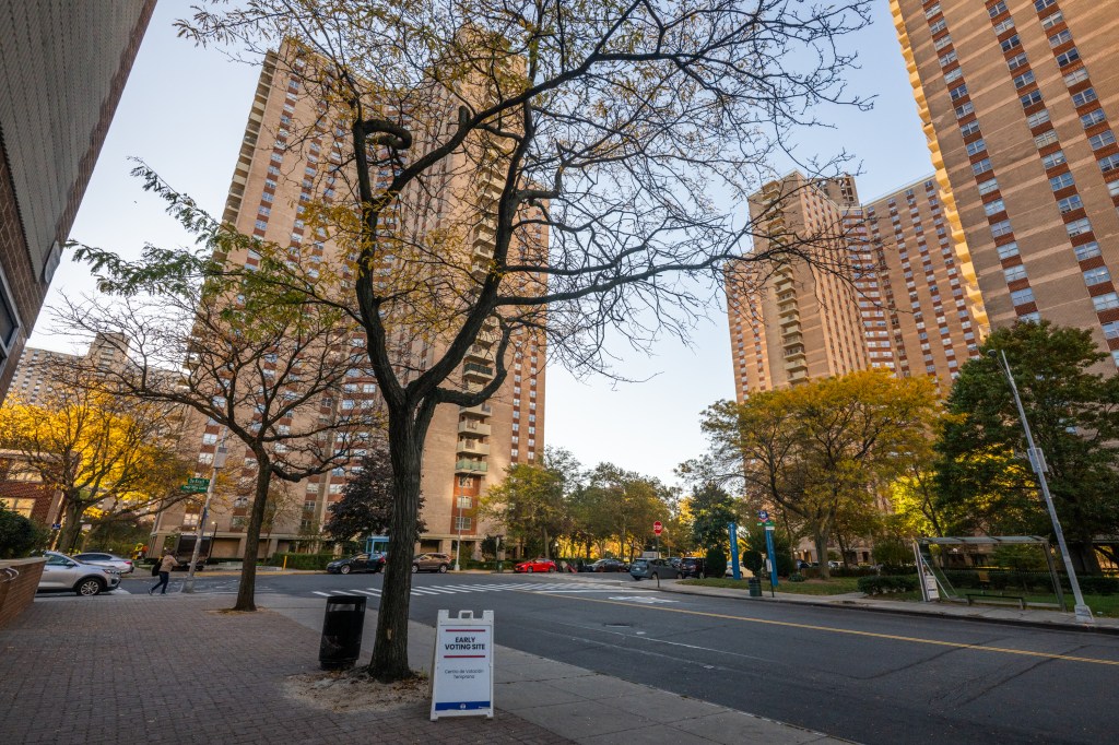 An "Early Voting Site" sign on a sidewalk next to a road, with tall apartment buildings in the background.