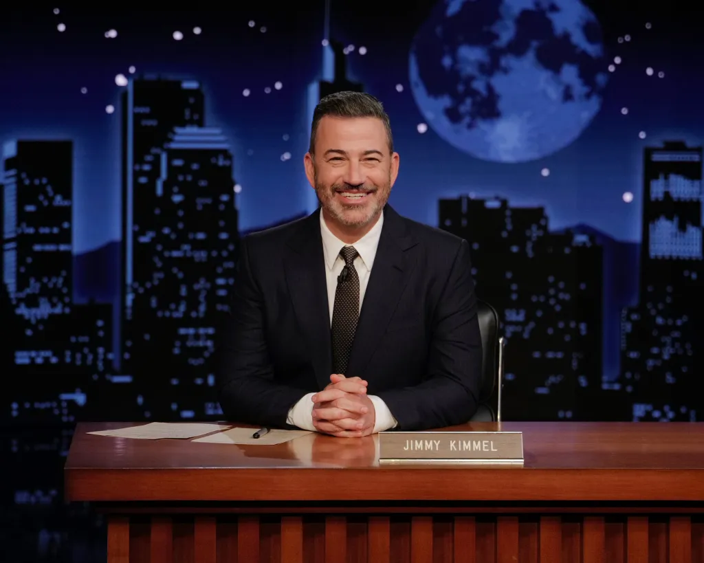 Jimmy Kimmel smiling at his desk with a cityscape and moon in the background.