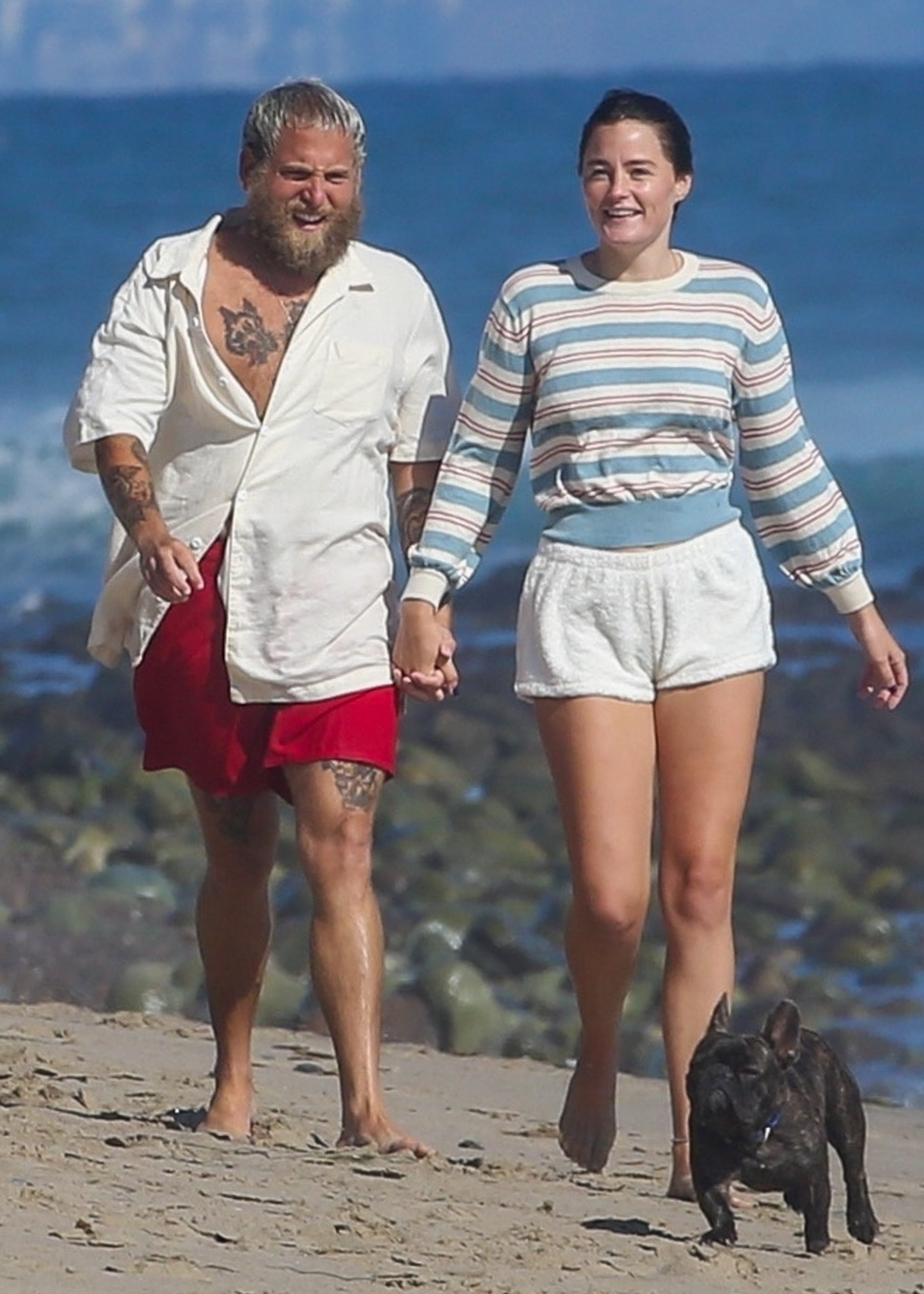 Jonah Hill and Olivia Millar walking hand-in-hand on the beach with a small dog.