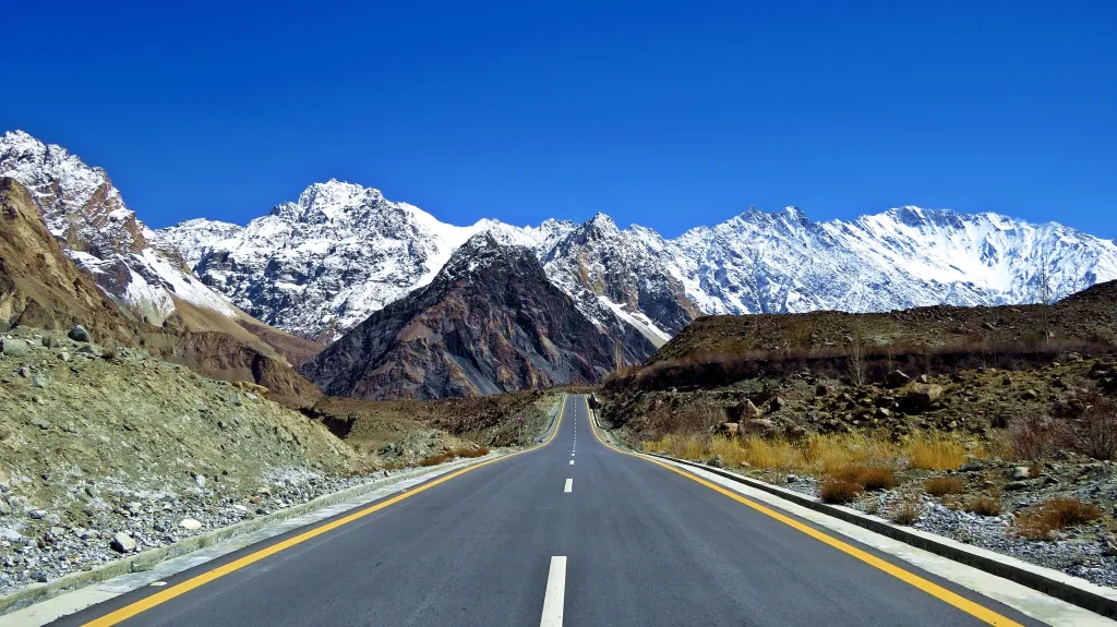 The Karakoram Highway, a high-altitude road in Pakistan, curving through a valley surrounded by snow-capped mountains under a clear blue sky.