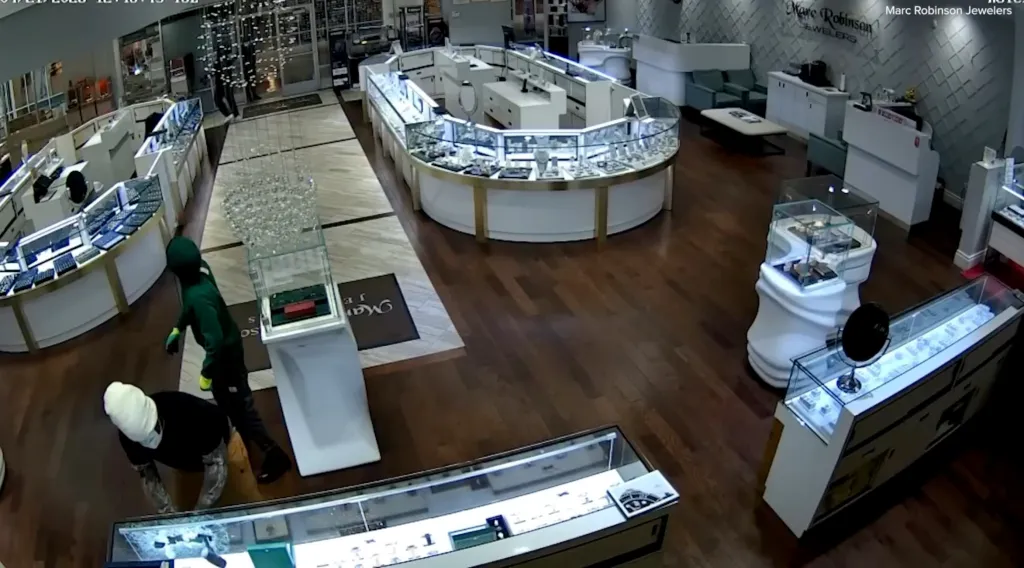 Two masked individuals in a jewelry store, one breaking a display case and the other standing nearby.