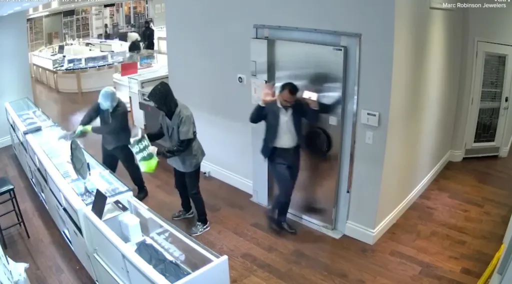 A security camera still of a jewelry store robbery in progress, with two masked individuals breaking a display case and holding bags, while a man in a suit backs away from a large metal safe.