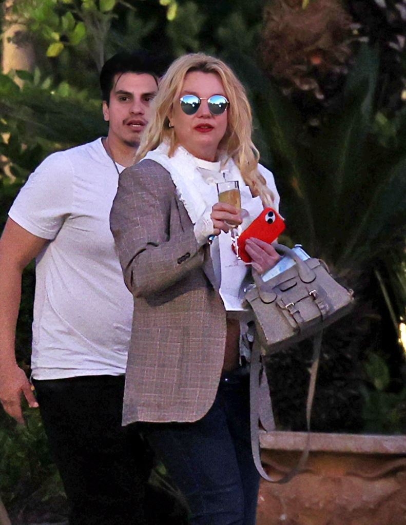 Britney Spears in sunglasses, holding a drink and a red phone, with a man behind her.