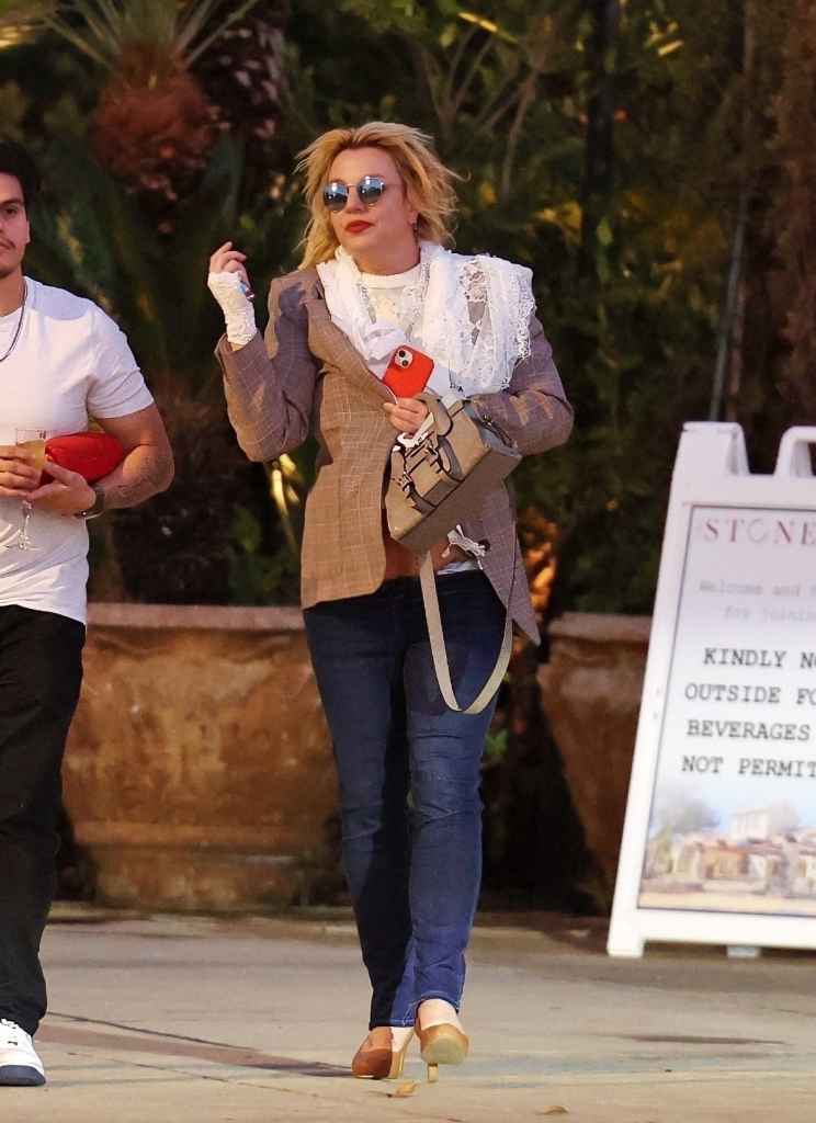 Britney Spears in sunglasses, brown blazer, and blue jeans, holding an orange cell phone and a light-colored handbag, walking next to a man.